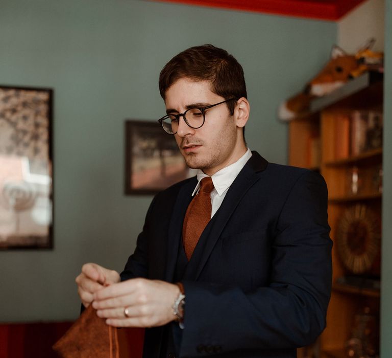 Groom in brown glasses wears blue suit with rust coloured tie and folds his matching handkerchief