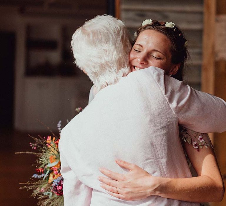 Bride with flowers in her hair hugs a wedding guest holding colourful wedding bouquet 