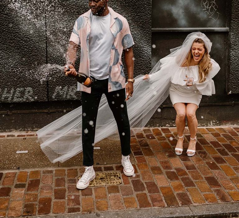 Groom in a pastel patterned shirt and Converse high tops popping champagne with his bride in a short wedding dress and long veil