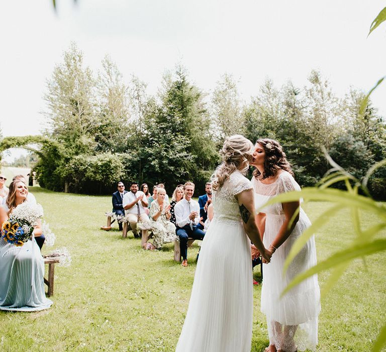 Brides share their first kiss as a married couple as wedding guests clap at outdoor ceremony