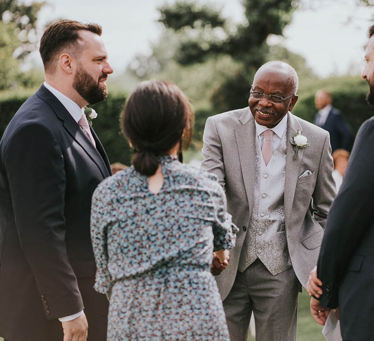 Father of the bride in light grey suit and patterned waistcoat and pink tie socialises with wedding guests