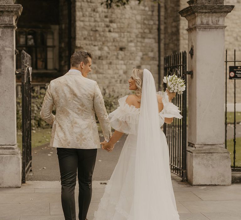 Bride in ruffle wedding dress walks with groom in custom tuxedo holding white tulip wedding bouquet