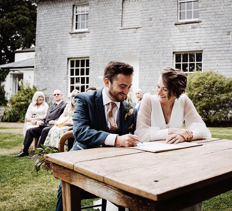 Groom in blue suit and gold patterned tie signs the register with the bride in Sassi Holford wedding gown