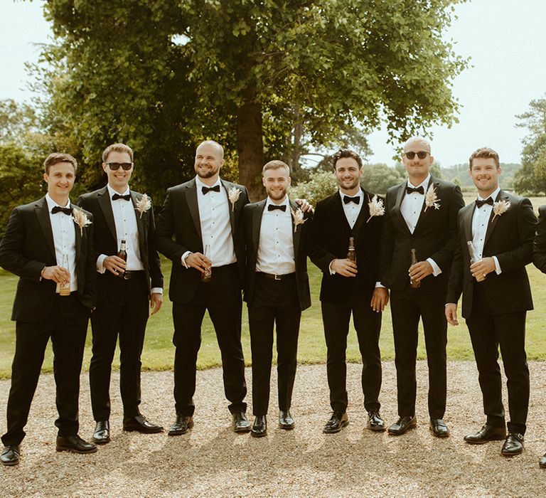 Groom with eight groomsmen in black tie and matching dried flower buttonholes drinking for summer wedding