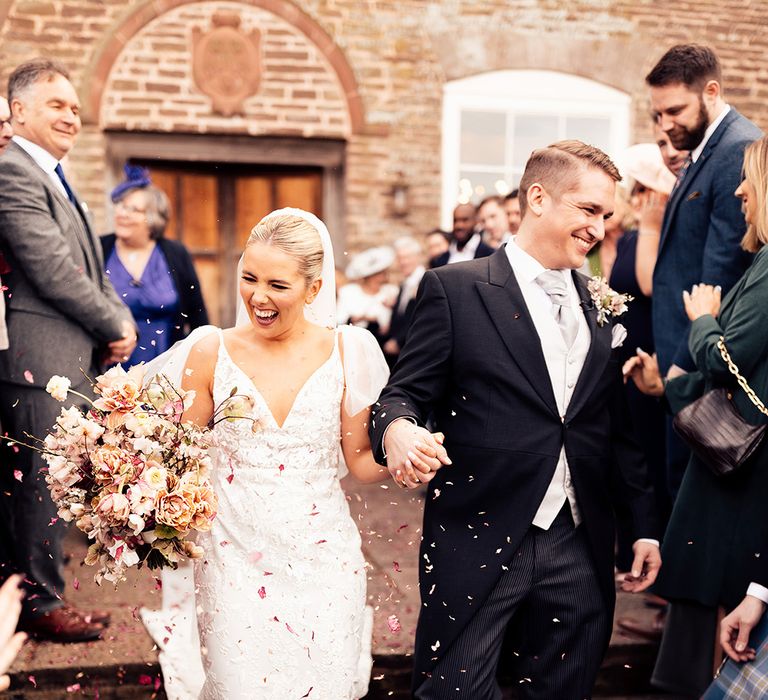 Bride in lace wedding dress with ruffle sleeves smiles at wedding guests as groom does the same in his morning suit with grey tie 