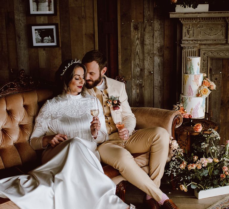 Bride in a satin wedding dress with fringe detail sitting on a sofa with her groom in a beige suit at Montague Farm Hankham rustic barn wedding venue 