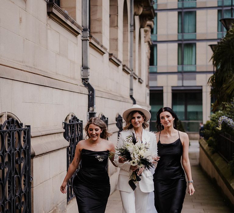 Bride wearing hat and pearl white trouser suit with bridesmaids in black satin dresses 