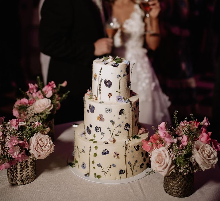 Three tier white frosted wedding cake with purple wildflowers and sprigs of lavender next to vases of pink flowers 