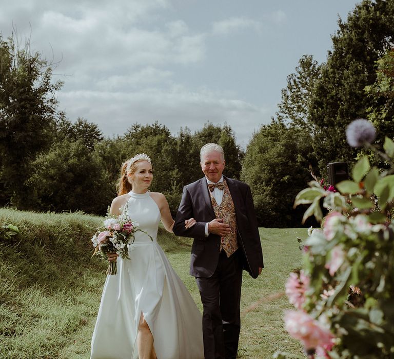 Bride in halter neck wedding dress wit pearl shoes is walked down the aisle by father of the bride in three piece suit with paisley tie and waistcoat 