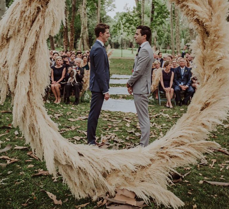 Two grooms stand for their wedding ceremony in front of a full pampas grass moongate 