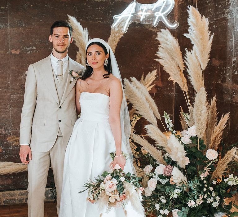 Pampas grass floral arrangement with personalised white neon sign with bride and groom standing in front 