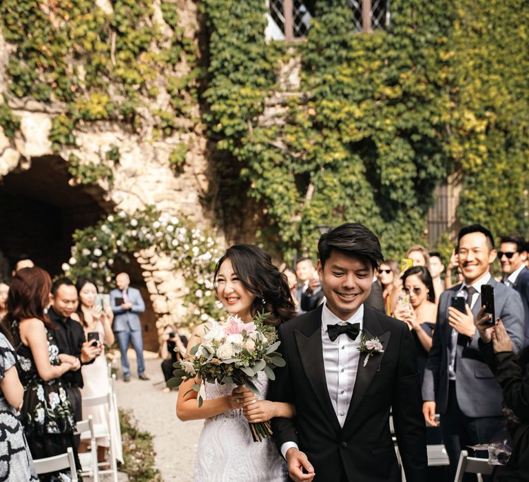 Groom wears black-tie complete with floral buttonhole as he walks down the aisle with his bride who wears lace embellished wedding dress whilst holding pastel floral bouquet