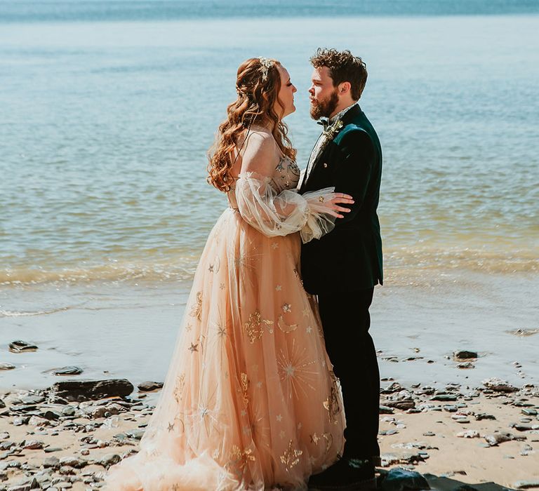 Bride wears off-the-shoulder starry wedding dress whilst stood with her groom in green velvet jacket on the beachfront in Cornwall 