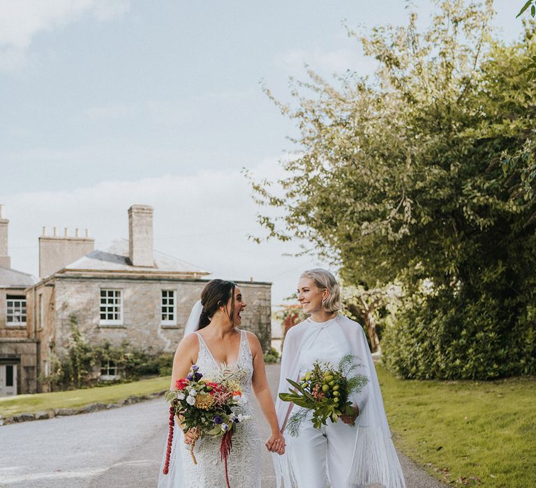 Bride in a handmade wedding jumpsuit and tassel cape walks holding hands with the bride in a lace wedding dress