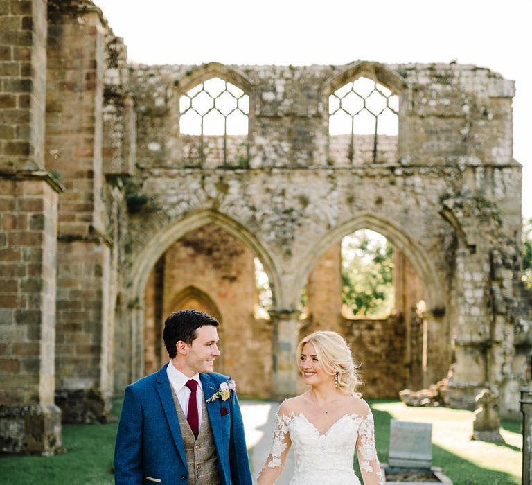 Bride wears long sleeve lace Pronovias wedding dress and holds her grooms hand as they walk through castle ruins on their wedding day