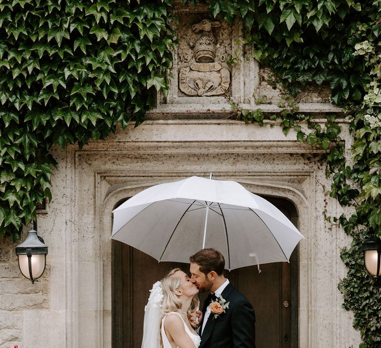 Bride in a floral applique wedding dress kisses the groom in a black tie under an umbrella 