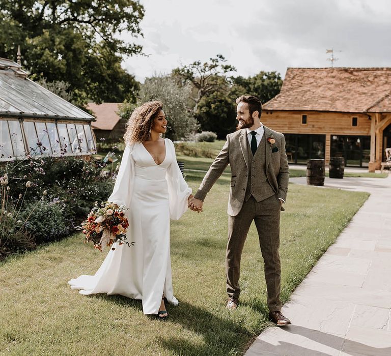 Bride in a satin wedding dress with balloon sleeves walking with the groom in a grey tweed suit 