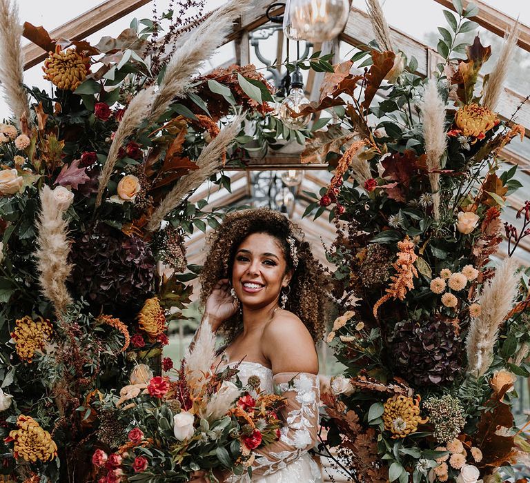 Bride in an off the shoulder wedding dress with pearl jewellery and accessories in front of autumnal flower columns with pampas grass