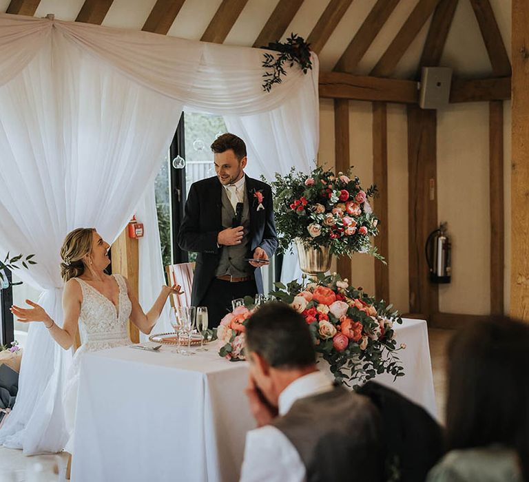 Groom stands to read out his speech as the bride sits at their sweetheart table decorated with pink flowers 