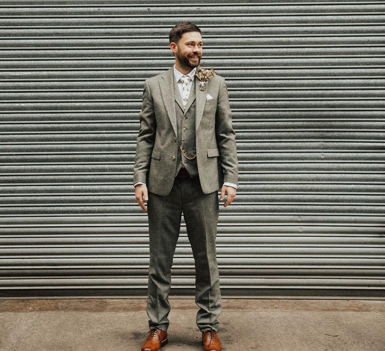 Groom wears three-piece grey suit with dried floral buttonhole and white pocket square 