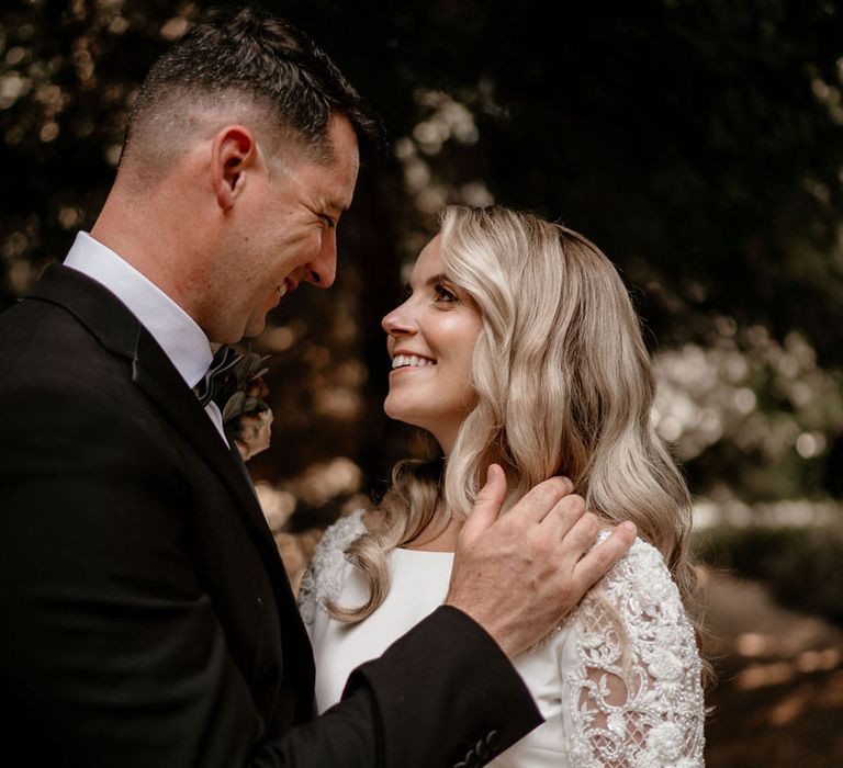 Bride with blonde curled hair in a beaded long sleeve wedding dress looking into the groom's eyes 