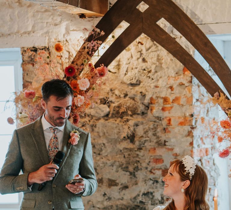 Groom in a tweed suit with a floral tie stands up and delivers his wedding speech as the bride smiles up at him 