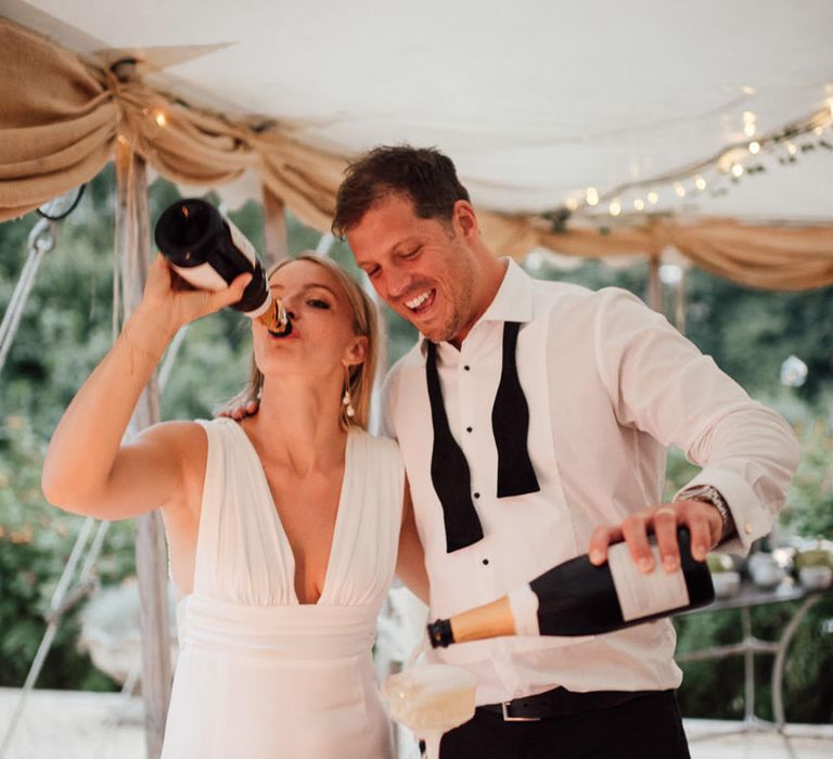 The bride and groom pour champagne for their champagne tower as the bride drinks from the bottle 