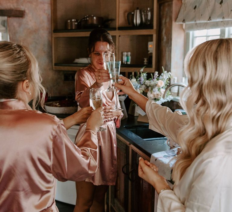 Bride in white satin robe shares a toast with the bridesmaids in pink satin robes as they get ready for the wedding 