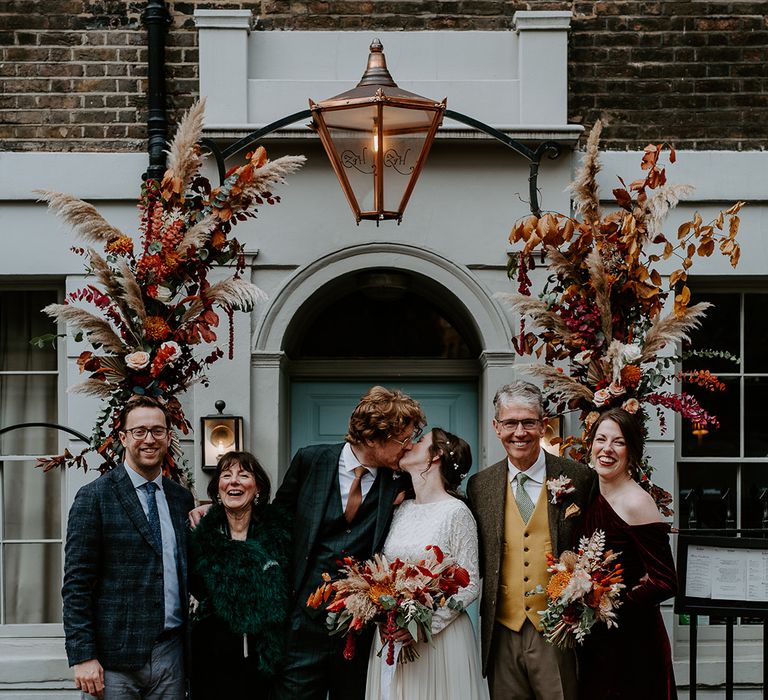 Bride in long sleeve lace vintage wedding dress and closed toe satin heels and groom in dark suit with orange tie and autumnal boutonniere kissing outside London townhouse with family of the bride and groom standing underneath large floral arch with roses, peonies, foliage and pampas grass