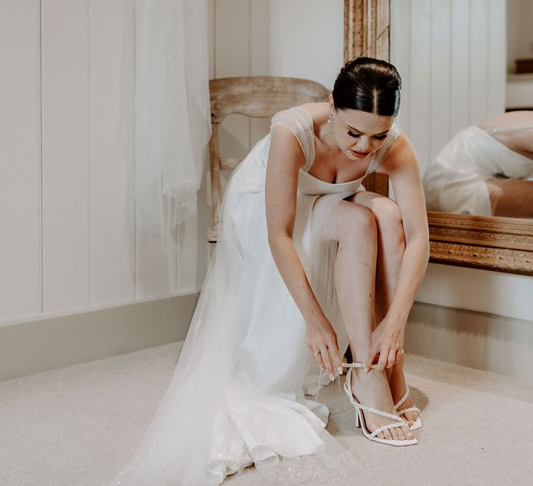 Bride wearing sparkly wedding dress puts on her white wedding shoes with DIY pearls 