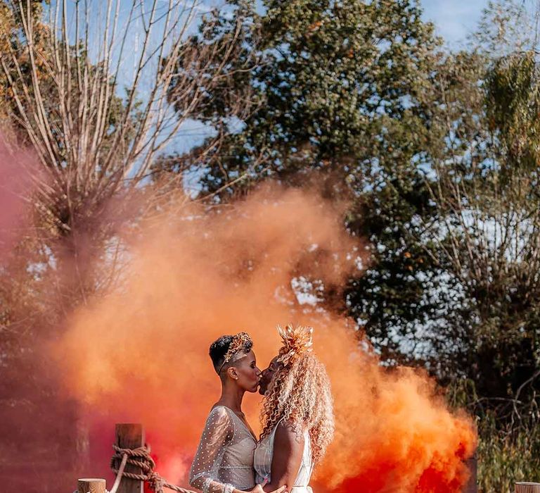 Brides in complimenting boho wedding dresses and golden bridal crowns kissing in front of large orange smoke bomb at Silchester Farm