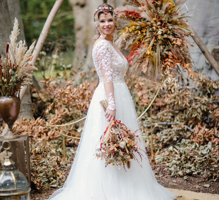 Bride in long sleeved v neck soft ivory dress with lace floral detailing on the bodice and sheer sleeves standing by large triangular wooden wedding arch with dried flower arrangements 