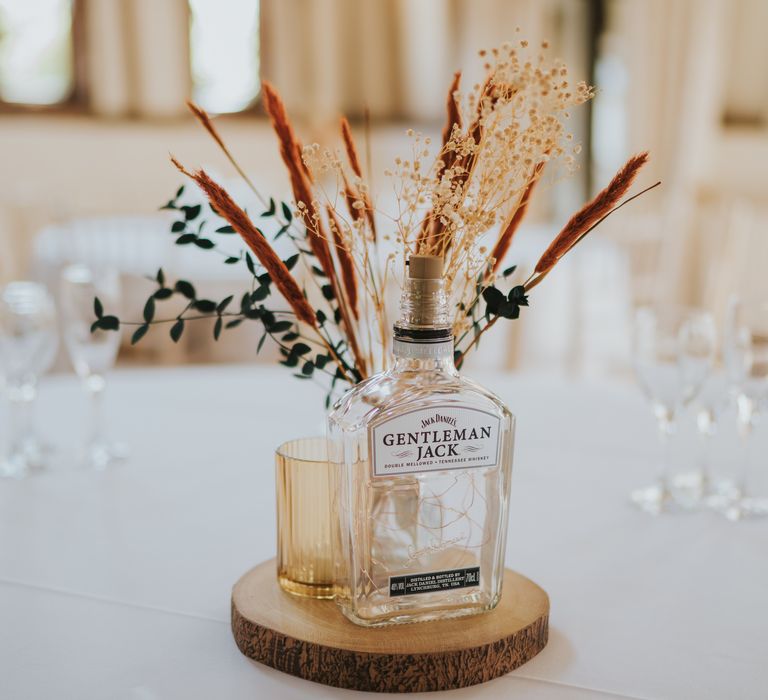 Wooden slate centrepiece on neutral wedding tablescape with clear alcohol bottle and clear vase with white bunny tails, green foliage and baby’s-breath