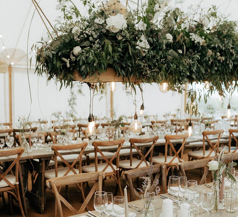 Marquee wedding reception with wooden banquet tables, green foliage hanging installations and fairy light canopy