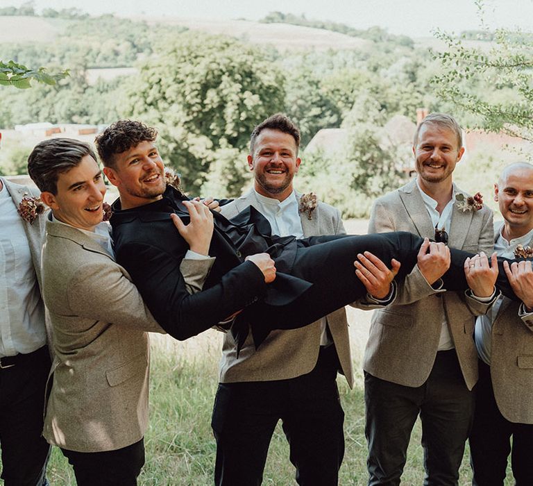 Groomsmen in beige holding up the groom in an all black suit with DIY dried buttonholes