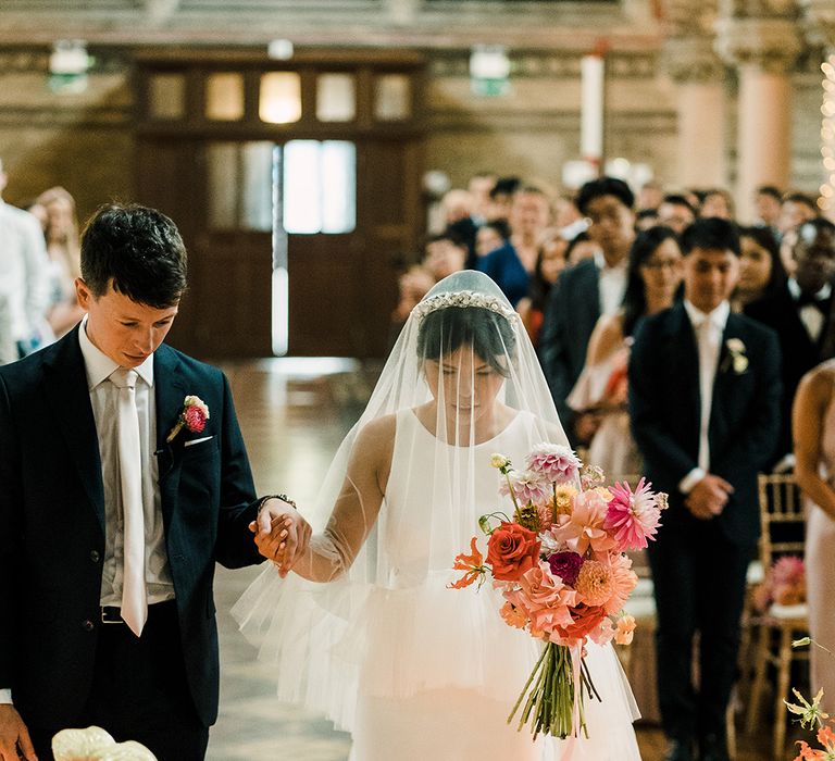 Bride holds colourful floral bouquet and wears drop ruffled veil whilst holding the hand of her groom during church wedding ceremony