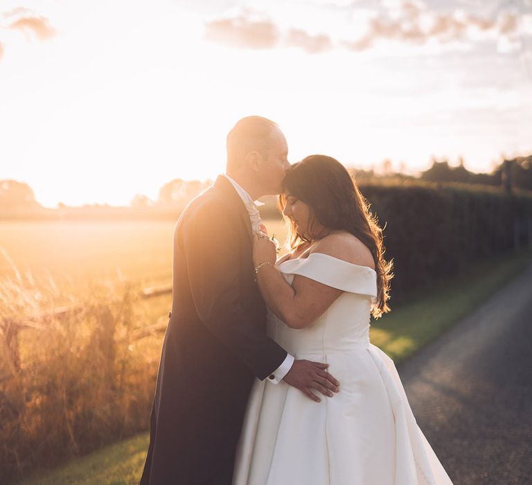 The groom in a morning suit kisses the bride on the forehead during sunset golden hour 