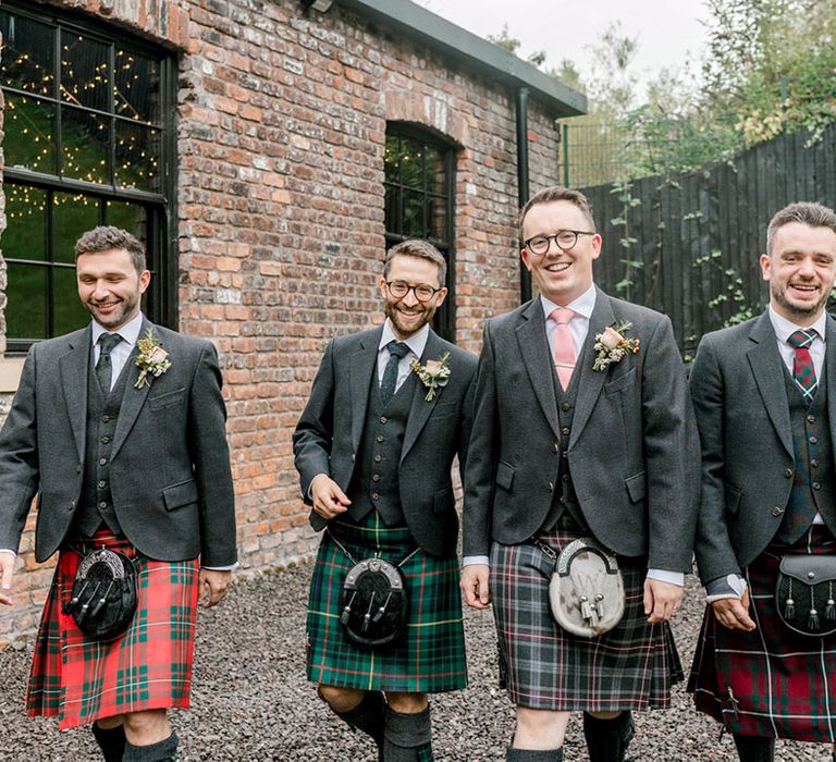 Groom walks alongside his groomsmen in different coloured tartan kilts and grey jackets with pastel ties 