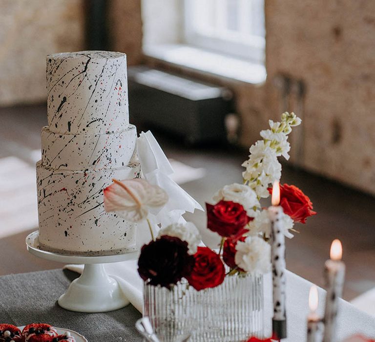 Black red and white wedding decor - three tier buttercream red velvet wedding cake with black paint splatter icing, and red and white roses, carnations and chrysanthemums, black and white tapered candles in clear candlestick holders