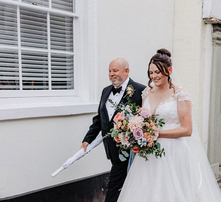 Bride in traditional princess skirt flower Suzanne Neville wedding dress with spring wedding bouquet walking with the father of the bride in black tie 