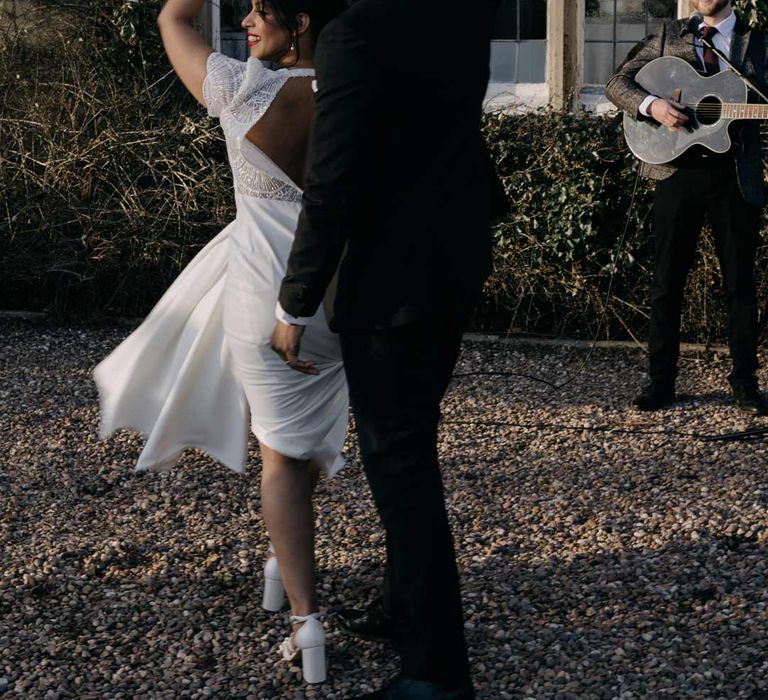 Bride in lace open back detail wedding dress with short sleeves and a large pearl headband dancing with groom in black tuxedo to live acoustic wedding band 