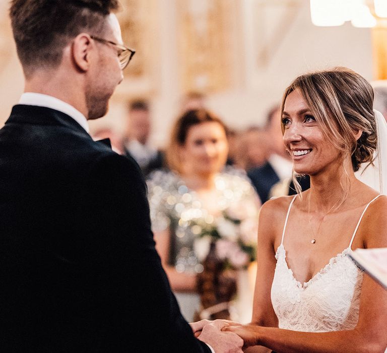 The bride wears a lace spaghetti strap wedding dress with her hair in an updo facing the groom for their church wedding ceremony