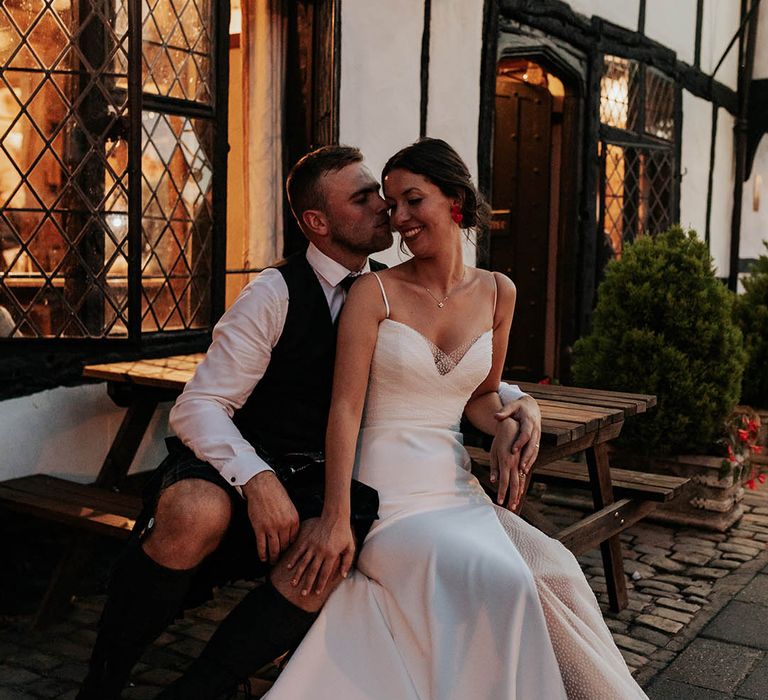 Bride in a Stephanie Allin bridal gown with thin straps, front slips and polka dot sheer overlay sitting on her grooms lap outside their wedding venue 