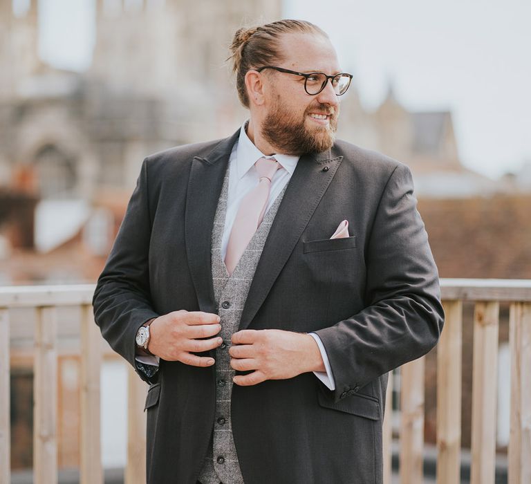 Groom in dark grey suit jacket and trousers with light grey checkered waistcoat and pastel pink tie and flower buttonhole 