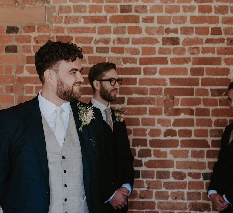 Groom in navy suit jacket with a light beige waistcoat and pale tie with a flower buttonhole turns to get his first look at the bride 