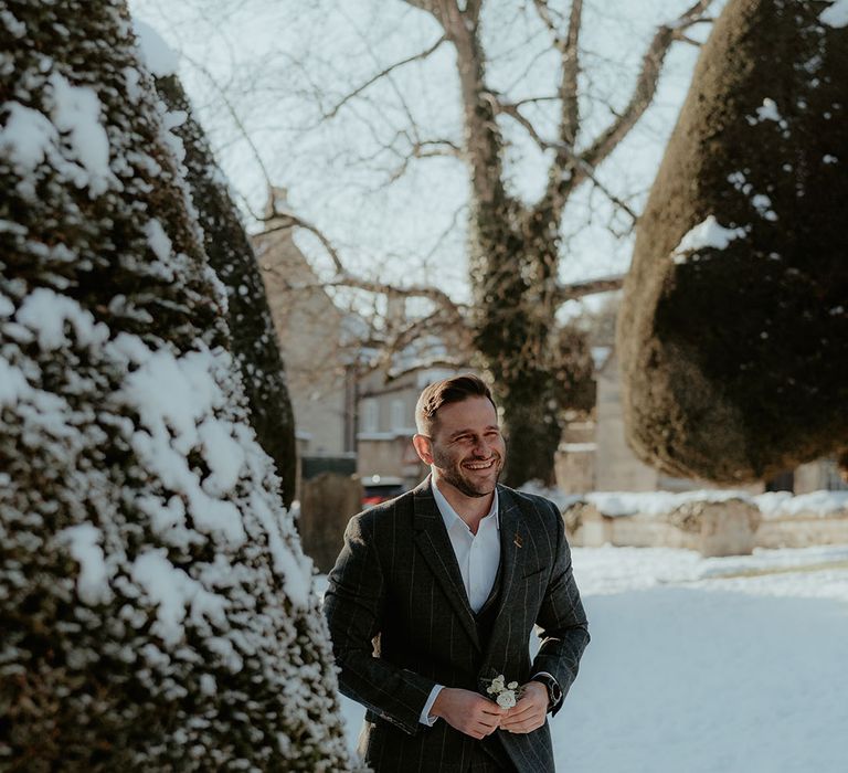 Groom in grey checkered suit walks through the snow in the Cotswolds to the bride for the first look moment 