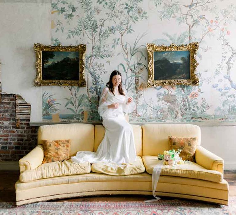 Bride sitting on top of yellow velvet sofa in reception room of St Giles House wearing cowl neck satin wedding dress with sheer puff sleeves