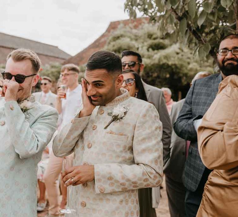 Groom in cream and gold Indian sherwani and white garden rose boutonniere standing with groom in mint green Indian Sherwani with white garden rose and foliage boutonniere and grooms sunglasses listening to wedding speeches at High Billinghurst Farm