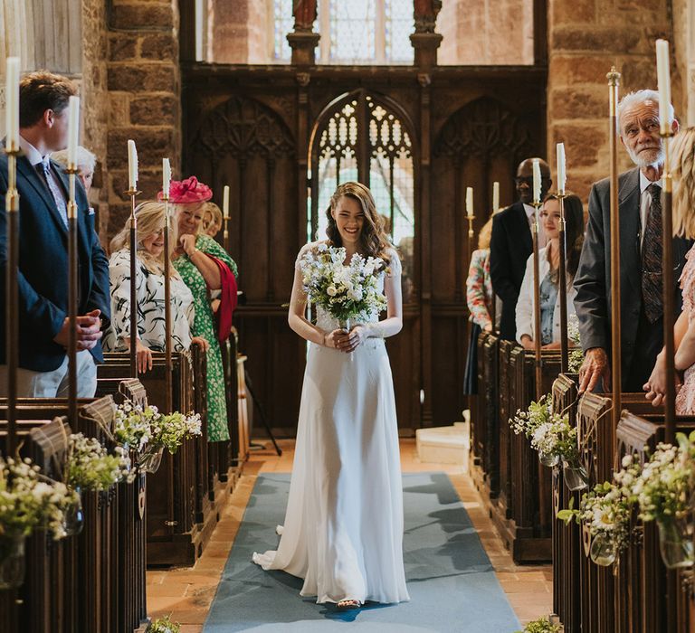 Bridesmaid with short sleeves and bejewelled detail walking down the aisle by herself for the Christian wedding 