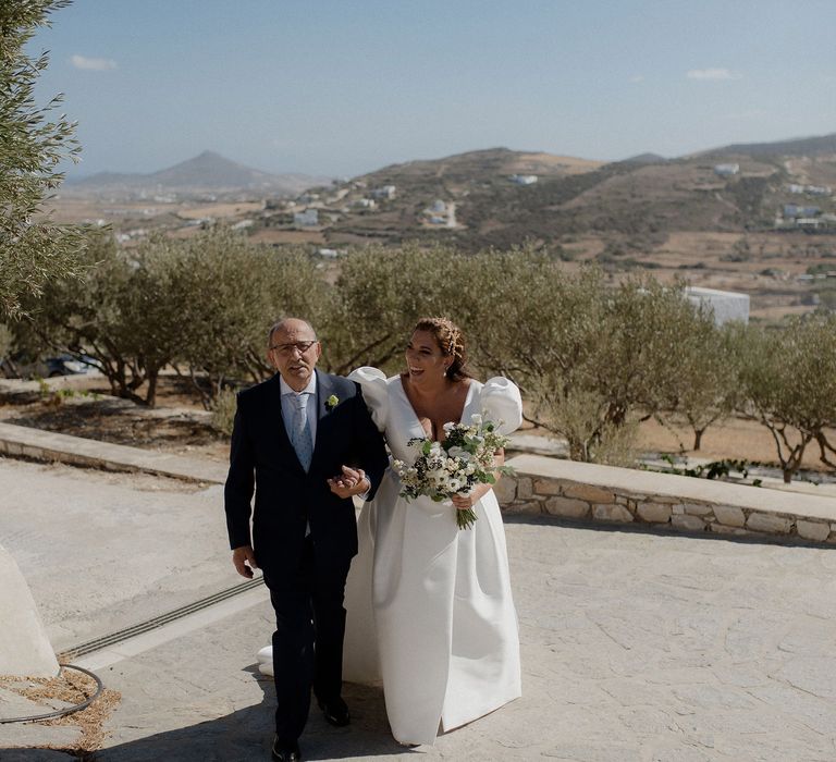 Father of the bride walks bride in satin white dress into wedding ceremony at destination wedding in Paros